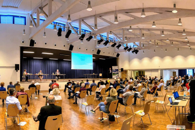 Saal mit Zuschauern beim Bürgerforum Energiewende Hessen. Auf der Bühne sitzen einige Personen an Tischen mit einer großen Videoleinwand im Hintergrund.